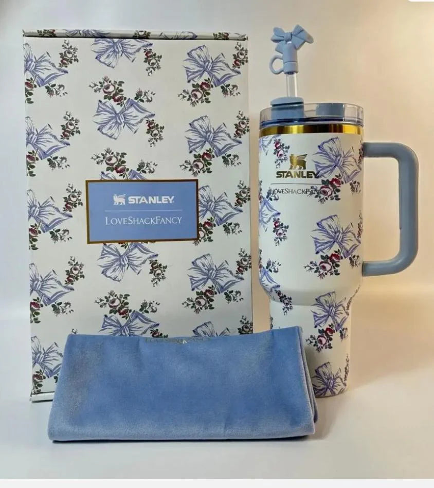 Stanley insulated travel mug with a floral and bow pattern, shown with its branded gift box and a folded blue cloth on a light beige background.