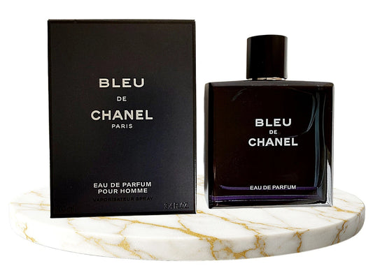Chanel Bleu de Chanel Eau de Parfum bottle and its black packaging box, displayed on a white marble round tray with gold veining against a white background.
